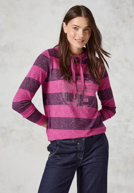 Frau im pink-lila gestreiften Pullover und Jeans, lächelnd vor hellem Hintergrund.